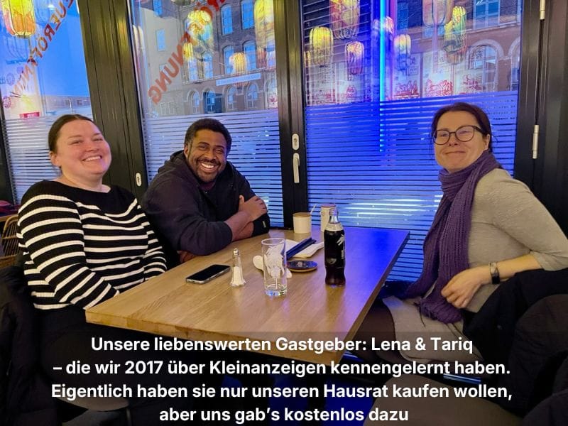 Andrea sitzt mit ihren Freunden Lena und Tariq an einem Vierertisch im Restaurant, alle lachen in die Kamera.