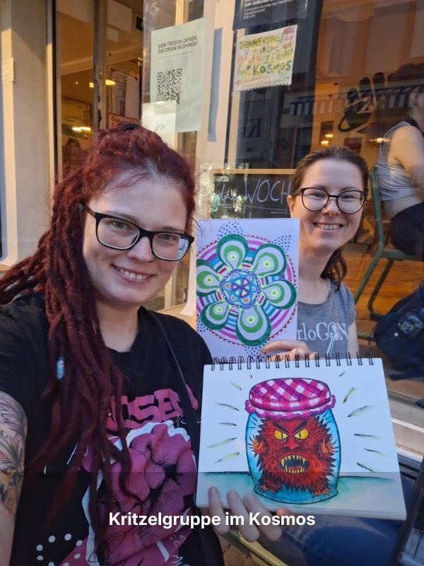 Andrea sitzt mit Tamara Leonhard vor einem Lokal im Freien, beide halten ihre selbstgemalten Bilder in die Kamera und lächeln.